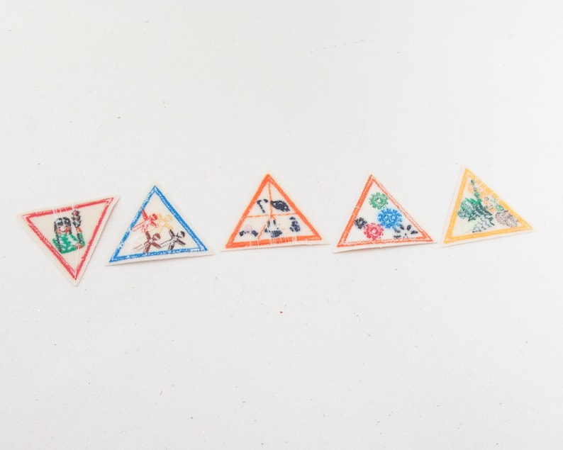 May include: Five triangular patches with various designs. The patches have a white background and are outlined in red, blue, orange, and yellow. The designs include floral patterns and abstract figures. These patches could be used for sewing or crafting projects.