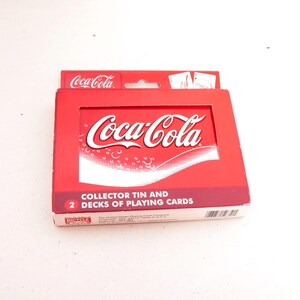 May include: A red Coca-Cola collector's tin containing two decks of playing cards. The tin features the Coca-Cola logo in white and the words "COLLECTOR TIN AND 2 DECKS OF PLAYING CARDS".