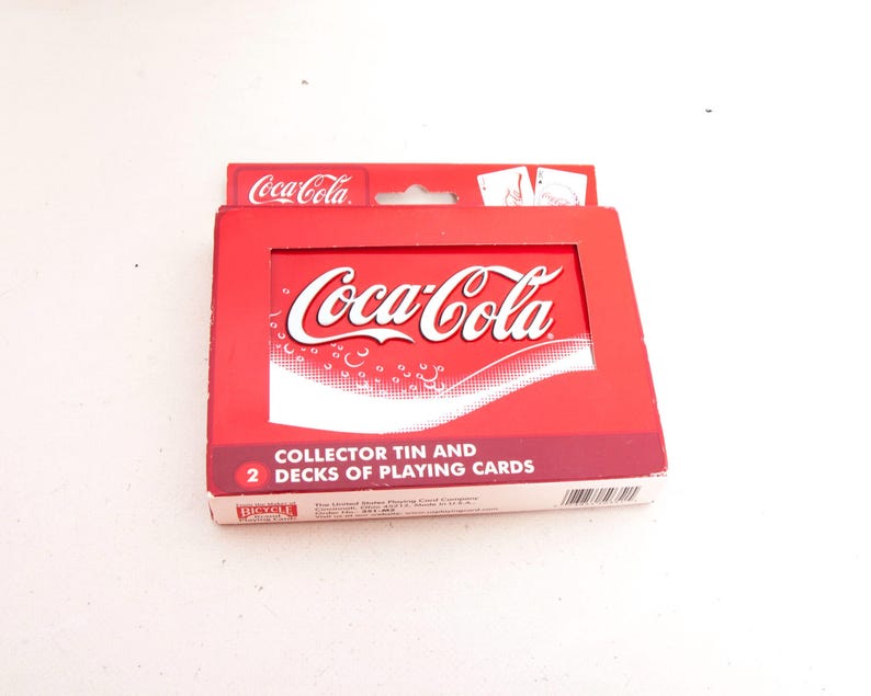 May include: A red Coca-Cola collector's tin containing two decks of playing cards. The tin features the Coca-Cola logo in white and the words "COLLECTOR TIN AND 2 DECKS OF PLAYING CARDS".