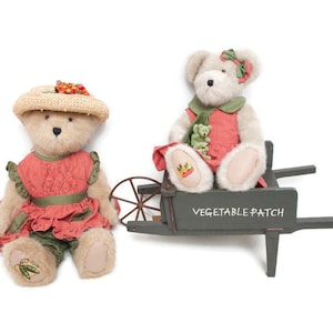 May include: Two plush teddy bears. One bear wears a straw hat and a red dress with green accents. The other bear sits in a gray wheelbarrow with the words "VEGETABLE PATCH" and wears a red dress and green scarf.