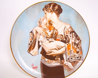 May include: A round decorative porcelain plate featuring a detailed illustration of a person holding a small child. The artwork uses a color palette of light blue, brown, orange, and white, finished with a thin gold rim around the edge.