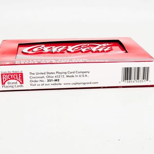 May include: A red and white Coca-Cola collector's tin containing two decks of Bicycle brand playing cards. The tin features the Coca-Cola logo and the Bicycle brand logo. The packaging includes text from The United States Playing Card Company.
