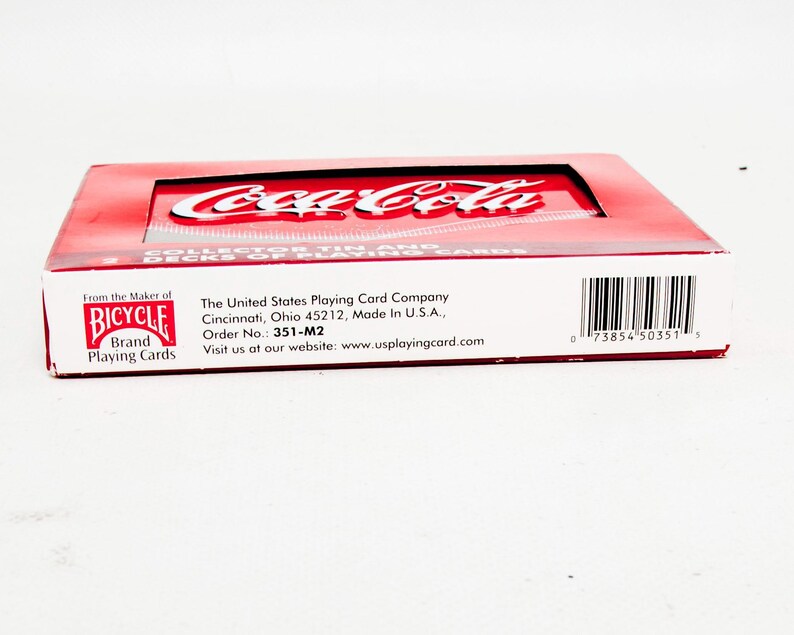 May include: A red and white Coca-Cola collector's tin containing two decks of Bicycle brand playing cards. The tin features the Coca-Cola logo and the Bicycle brand logo. The packaging includes text from The United States Playing Card Company.