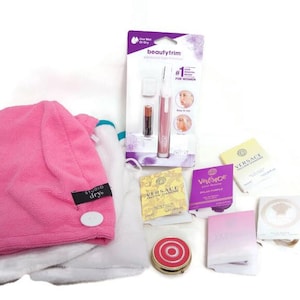 Puede incluir: Una colección de artículos de belleza y cuidado personal, que incluye una toalla rosa para el cabello, una herramienta de depilación beautytrim, muestras de perfume y un pequeño espejo compacto.