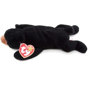 May include: A black plush bear toy with a brown snout and a red and white heart-shaped tag. The bear is lying down on a white surface.