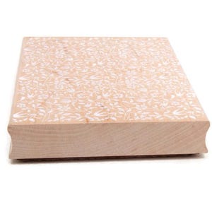 Puede incluir: Un sello cuadrado de madera con un diseño floral. El sello está hecho de madera de color claro y presenta un diseño repetido de flores y hojas blancas. Los bordes están ligeramente redondeados.