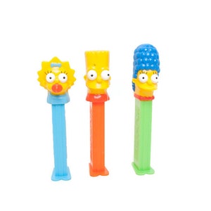 May include: Three PEZ dispensers featuring characters from The Simpsons. Maggie Simpson has a yellow head and blue stem. Bart Simpson has a yellow head and orange stem. Marge Simpson has a blue head and green stem.