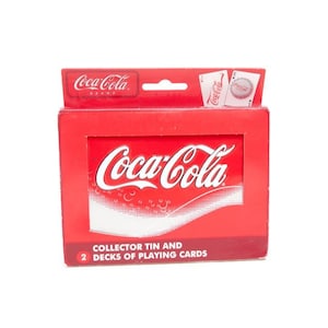 May include: A red Coca-Cola collector's tin box with the Coca-Cola logo in white. The box contains two decks of playing cards. The top of the box has the Coca-Cola brand logo and two playing cards.
