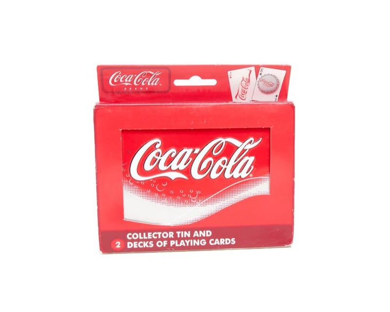 May include: A red Coca-Cola collector's tin box with the Coca-Cola logo in white. The box contains two decks of playing cards. The top of the box has the Coca-Cola brand logo and two playing cards.