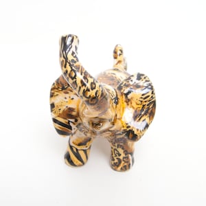 Könnte beinhalten: Eine Keramik-Elefantenfigur mit einem einzigartigen Tiermuster. Der Körper des Elefanten weist eine Mischung aus braunen, beigen und schwarzen Mustern auf, die einem Leoparden oder Geparden ähneln. Der Rüssel ist erhoben und die Ohren sind ausgebreitet, was die detaillierte Handwerkskunst zeigt.