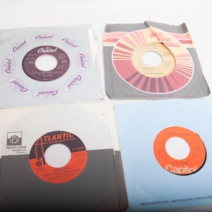 May include: Four vintage vinyl records in their sleeves. The records are in various colors, including red, orange, and purple. The sleeves feature the Capitol and Atlantic record labels. The records are arranged on a white surface.