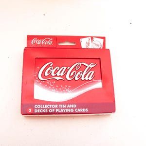 May include: A red Coca-Cola branded collector's tin containing two decks of playing cards. The tin features the Coca-Cola logo in white script on a red background. The text "COLLECTOR TIN AND 2 DECKS OF PLAYING CARDS" is printed on the bottom of the tin.