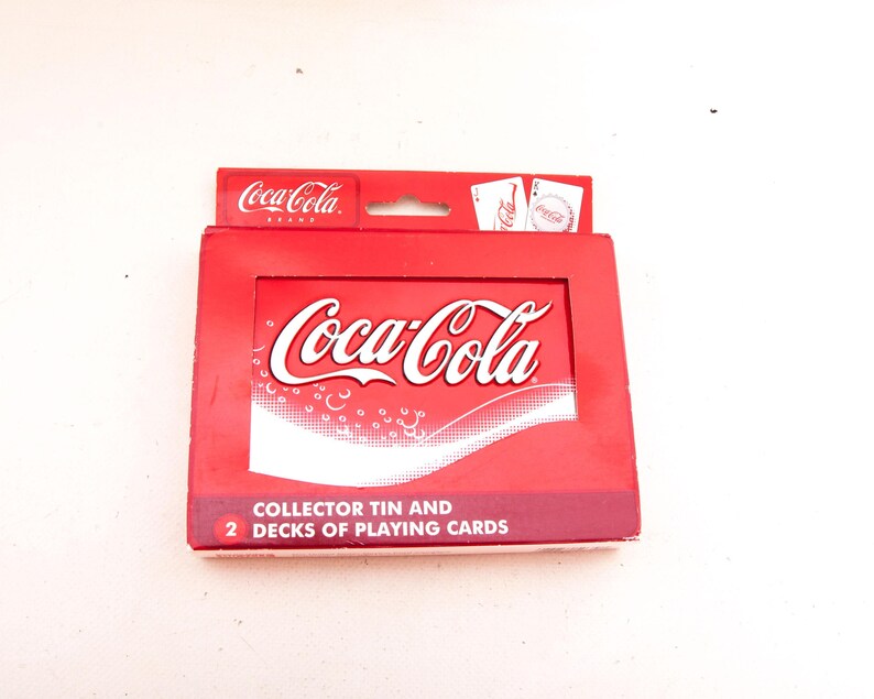 May include: A red Coca-Cola branded collector's tin containing two decks of playing cards. The tin features the Coca-Cola logo in white script on a red background. The text "COLLECTOR TIN AND 2 DECKS OF PLAYING CARDS" is printed on the bottom of the tin.