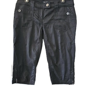 May include: Black capri trousers with multiple pockets and decorative buttons. The trousers are hanging on a black hanger. The capris have zips at the bottom of each leg.
