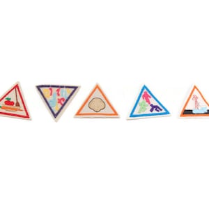 May include: A collection of five triangular patches with embroidered designs. The patches feature images of food, musical notes, a cookie, figures walking, and a person taking a bath. Each patch has a white background with a colored border.