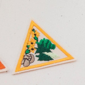 May include: Embroidered triangular patch with a yellow border and a cream-colored background. The patch features a green tree, yellow sunflowers, and brown mushrooms. The patch is likely for a scout or similar organization.