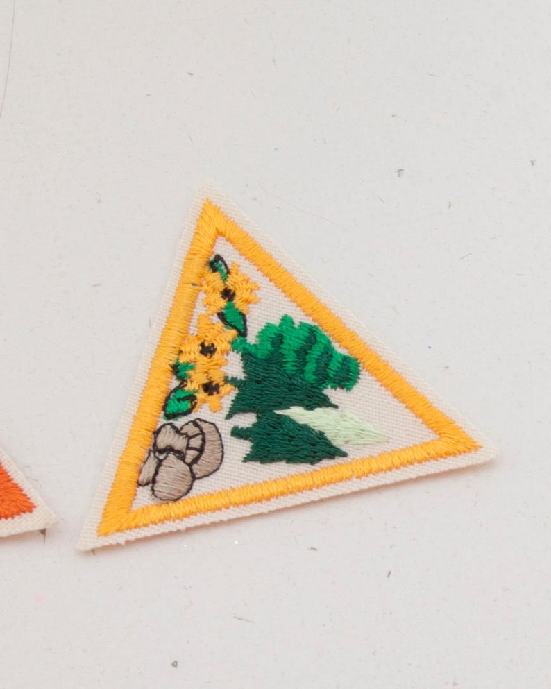 May include: Embroidered triangular patch with a yellow border and a cream-colored background. The patch features a green tree, yellow sunflowers, and brown mushrooms. The patch is likely for a scout or similar organization.