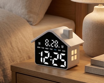 May include: A white house-shaped digital alarm clock with a black display screen showing the time 12:25, the date 8.28, and the days of the week. The clock features a small chimney, a side window with a warm light, and white digital numerals.