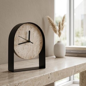 May include: A modern, minimalist clock with a circular face made of speckled beige stone. The clock is set in a black metal frame. The hands are black. The clock sits on a beige stone surface, with a vase and books in the background.