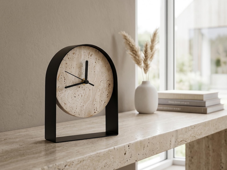 May include: A modern, minimalist clock with a circular face made of speckled beige stone. The clock is set in a black metal frame. The hands are black. The clock sits on a beige stone surface, with a vase and books in the background.