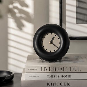 May include: A black, circular analog clock with a cream-coloured face and black hands. The clock sits atop a stack of three books with the titles "LIVE BEAUTIFUL", "THIS IS HOME", and "KINFOLK". The clock's diameter is approximately 10 cm.