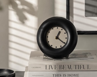May include: A black, circular analog clock with a cream-colored face and black hands. The clock sits atop a stack of three books with the titles "LIVE BEAUTIFUL", "THIS IS HOME", and "KINFOLK". The clock's diameter is approximately 4 inches.