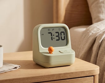 May include: A sage green digital alarm clock with a rounded rectangular frame and a monochrome LCD screen displaying 7:30 AM. The device features a prominent orange spherical button on the front left and a square power button on the front right.
