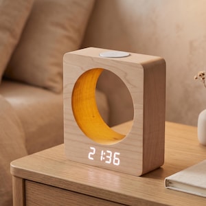 May include: A light-colored wooden digital clock with a square frame and a large circular cutout in the center. The interior of the circle features a warm yellow glow. A digital display at the bottom shows the time 21:36 in white light.