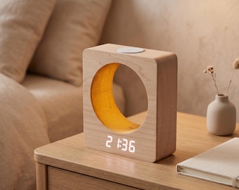 May include: A light-colored wooden digital clock with a square frame and a large circular cutout in the center. The interior of the circle features a warm yellow glow. A digital display at the bottom shows the time 21:36 in white light.