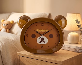 May include: A brown, bear-shaped analog clock with a glossy finish and two rounded ears. The clock face features the text Rilakkuma, black hour and minute hands, a red second hand, and a stylized bear face graphic with dark brown circular cheeks.