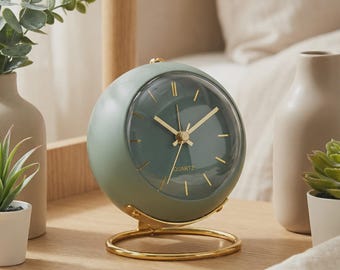 May include: A spherical sage green quartz table clock with a clear glass face, gold-toned hands, and gold-toned hour markers. The clock is mounted on a circular gold-toned metal stand, featuring the word QUARTZ printed on the clock face.