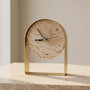 May include: A modern, gold-toned desk clock with a circular face featuring a marbled, beige pattern. The clock has black hands and a rectangular base. The clock is approximately 7 inches tall and 6 inches wide.