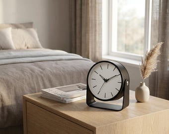 May include: A round analog table clock with a matte black frame and a white face featuring black hour markers and thin black hands. The clock is mounted on a U-shaped black metal stand that allows for tilting, positioned on a light wood surface.