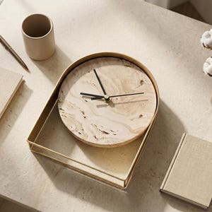 May include: A beige marble-faced clock with black hands sits in a gold-colored tray. A pen, a beige mug, and two beige books are on the table. A small glass vase with cotton flowers is also present. The scene is lit by natural light.