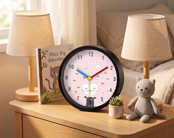 May include: A round wall clock with a black frame and a light pink face featuring a white cat illustration, small fish motifs, and black numerals. The clock has a blue hour hand, a red minute hand, and a yellow second hand centered on the dial.