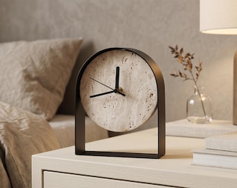 May include: A modern, minimalist clock with a circular face made of speckled stone. The clock has black hands and a black metal frame. It sits on a white nightstand, suggesting a bedroom setting. The clock's design is simple and elegant.