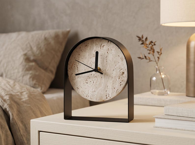 May include: A modern, minimalist clock with a circular face made of speckled stone. The clock has black hands and a black metal frame. It sits on a white nightstand, suggesting a bedroom setting. The clock's design is simple and elegant.