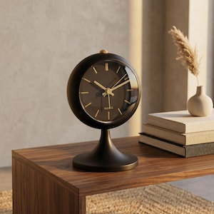 May include: A black quartz clock with gold hands and markers on a wooden table. The clock has a round face and a pedestal base. A small vase with a dried plant and books are also on the table.