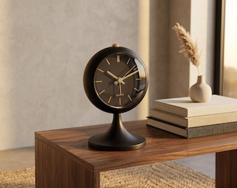 May include: A black quartz clock with gold hands and markers sits on a wooden table. The clock has a round face and a pedestal base. A small vase with a dried plant and a stack of books are also on the table.