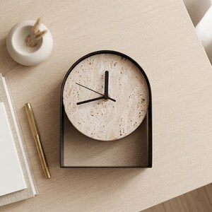 May include: A modern, minimalist clock with a speckled, light-colored face and black hands, set in a black metal frame. A gold pen, a small white vase with dried plants, and a stack of books are also on the light-colored wooden surface.