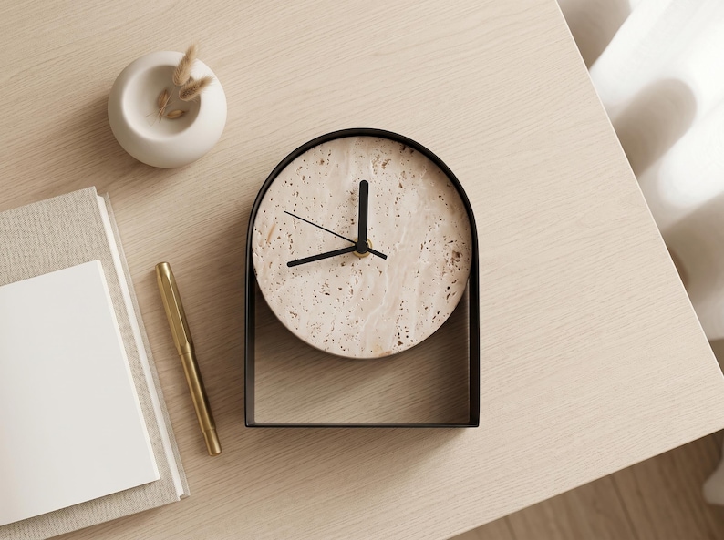 May include: A modern, minimalist clock with a speckled, light-colored face and black hands, set in a black metal frame. A gold pen, a small white vase with dried plants, and a stack of books are also on the light-colored wooden surface.