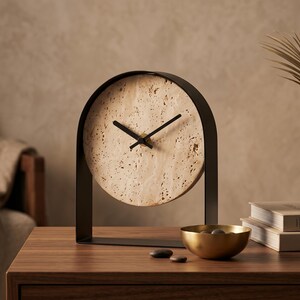 May include: A modern, minimalist table clock with a circular face made of speckled beige stone. The clock is set in a black metal arch frame. The hands are black. A gold bowl with stones and books are on the wooden table.