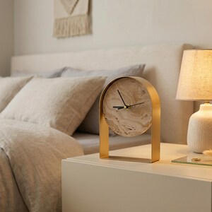 May include: A gold-colored arched desk clock with a marble-patterned face sits on a white nightstand next to a lamp with a textured white base and a beige lampshade. The clock's hands are black.