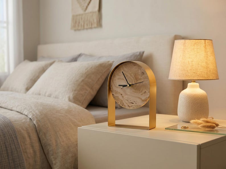 May include: A gold-colored arched desk clock with a marble-patterned face sits on a white nightstand next to a lamp with a textured white base and a beige lampshade. The clock's hands are black.