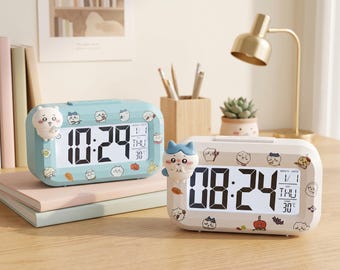 May include: Two rectangular digital alarm clocks, one light blue and one white, featuring rounded corners and cartoon character accents. Each clock displays the time, date, day of the week, and temperature on a clear LCD screen with black digits.