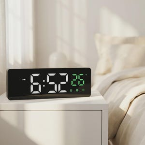 May include: A black digital alarm clock displaying the time 6:52 PM and the temperature 26 degrees. The clock sits on a white nightstand next to a bed with beige bedding. The clock has a rectangular shape.