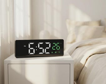 May include: A black digital alarm clock displaying the time 6:52 PM and the temperature 26 degrees. The clock sits on a white nightstand next to a bed with beige bedding. The clock has a rectangular shape.