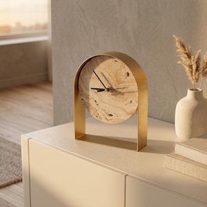 May include: A modern, gold-framed clock with a light-colored, patterned face sits on a white surface. The clock's hands are black. A small vase with dried plants and two books are next to the clock. The scene is lit by warm sunlight.