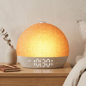 May include: A dome-shaped alarm clock with a warm, glowing orange fabric upper section and a light grey fabric base. The digital display shows the time 10:30 AM in white light, along with small icons for a snooze function, a musical note, and an alarm.