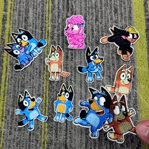 May include: A selection of embroidered patches depicting cartoon dog characters in different poses. The patches are in blue, brown, pink, and black, with white borders. The patches are displayed on a grey surface.